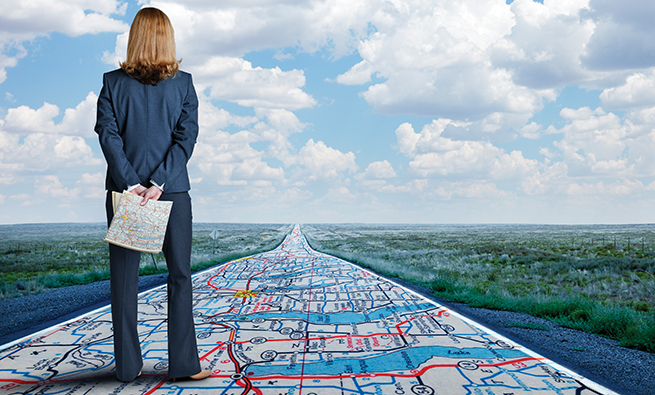 A businesswoman stands while holding a road map in her hands as she looks down a long, straight, rural road that has the same road map painted on it.